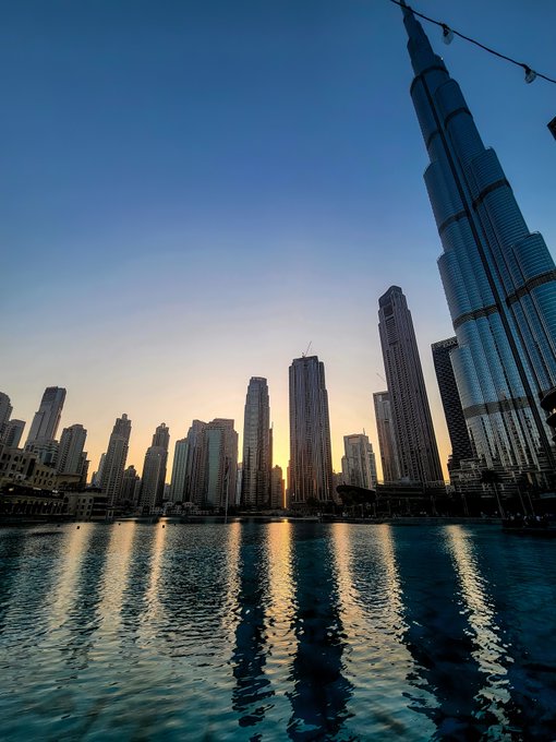A stunning sunset view of the Dubai skyline, featuring the iconic Burj Khalifa towering above the city with its skyscrapers reflecting in the calm waters below. The image celebrates the post's excitement about a successful secondary sale of a Crawford-curated Dubai NFT collection.