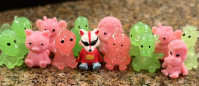 A collection of small, translucent pink and green animal figurines, including pigs, hedgehogs, and a fox-like character in a red outfit, arranged on a speckled surface.