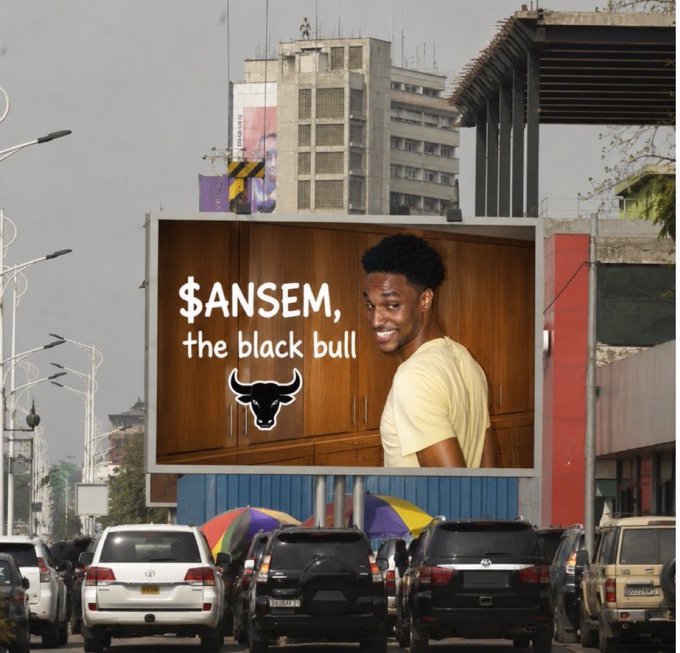 A billboard in a city street advertising '$ANSEM, the black bull' featuring a man in a yellow shirt alongside a Chicago Bulls-style logo.