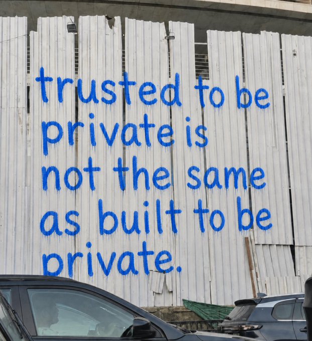 Blue spray-painted graffiti on a white wooden fence reads, 'trusted to be private is not the same as built to be private,' with parked cars visible at the bottom.