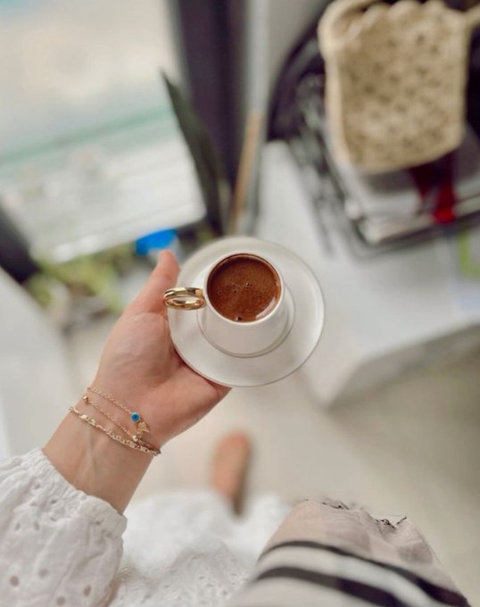 A person wearing delicate gold bracelets with a blue evil eye charm holds a small white cup of Turkish coffee, with a blurred home interior in the background. The cozy Sunday morning moment pairs with the post's relaxed tone, contrasting the heat and cleaning chores mentioned with a peaceful pause for coffee.