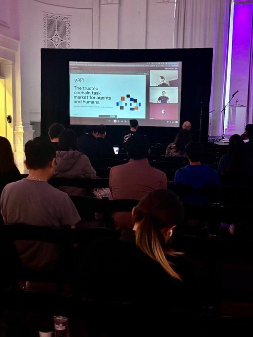 Audience at a conference viewing a presentation titled 'API: The trusted onchain task market for agents and humans' with video call participants displayed on a large screen.