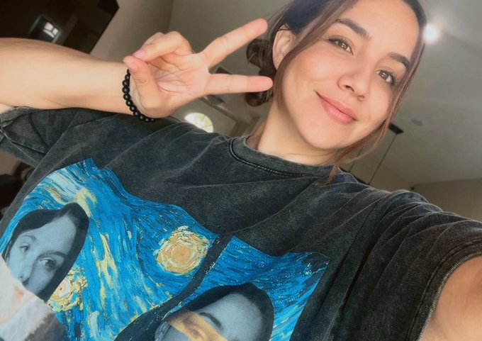 A woman smiles and flashes a peace sign in a selfie, wearing a vintage-style Van Gogh "Starry Night" graphic t-shirt, capturing a casual, upbeat moment. The relaxed pose and chill vibe match the "GM" (good morning) energy of her post about taking a step back to focus on building and personal growth.