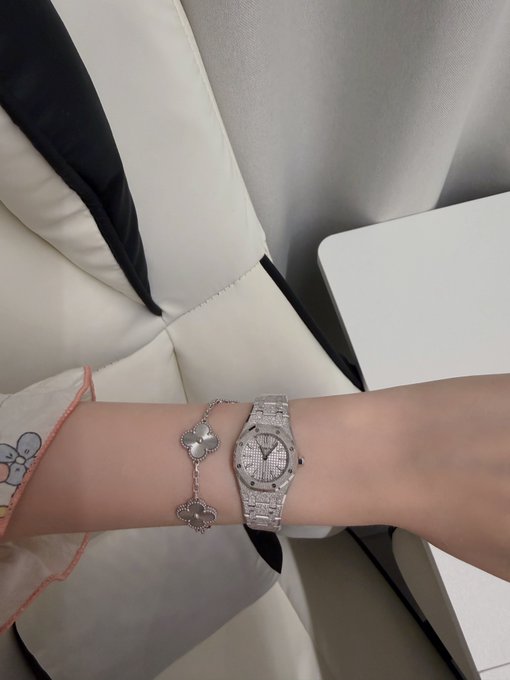 A wrist displaying a sparkly, diamond-encrusted Audemars Piguet watch alongside a four-leaf clover bracelet, captioned as a "mini frosted AP" gifted thanks to crypto and @ProsperityTime_.
