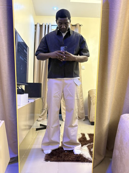 A young man takes a mirror selfie in a bedroom, wearing a black button-up shirt, cream-colored pants with a visible tag, and white socks, while holding a smartphone to capture the reflection. The "🦅❤️" caption suggests pride or affection, possibly tied to the new outfit or a personal milestone reflected in his confident pose.