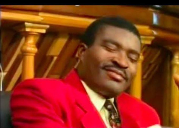 A man in a red suit and patterned tie sits with his eyes closed and a serene smile, set against ornate wooden background paneling. The image contrasts with the post's casual, humorous misspelled text about claiming free money online, humorously pairing a moment of peaceful composure with the chaotic, scammy-sounding idea of easy online cash.