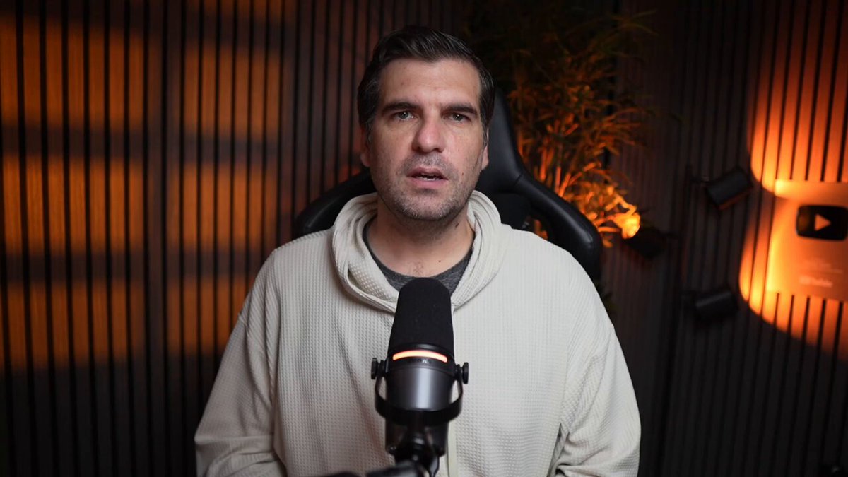 The image features a man wearing a white sweater standing behind a microphone, giving him an air of authority or expertise as he speaks into it. The background includes a dark wall with a plant located near the top left corner, adding some greenery to the scene. The main focus is on the man and his interaction with the microphone, which suggests that he might be discussing a topic related to smart contract verification, developer tooling, or familiar EVM workflows.