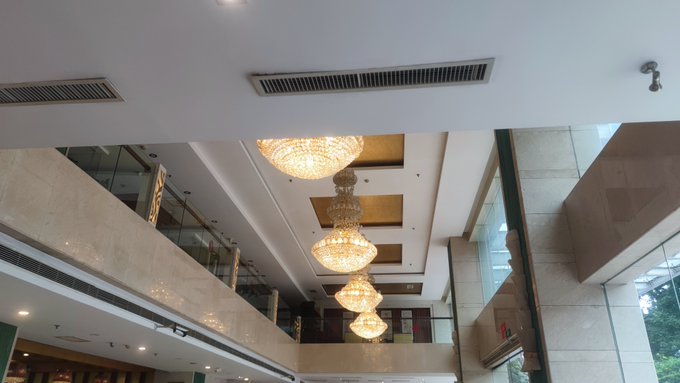 An interior view of a multi-story building lobby or atrium featuring ornate, large golden chandeliers hanging from a high ceiling, with marble walls and a balcony visible on an upper floor.