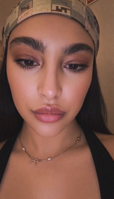 A close-up selfie shows a woman wearing a patterned headband and a delicate gold necklace, featuring prominent dark eyebrows and glossy makeup.