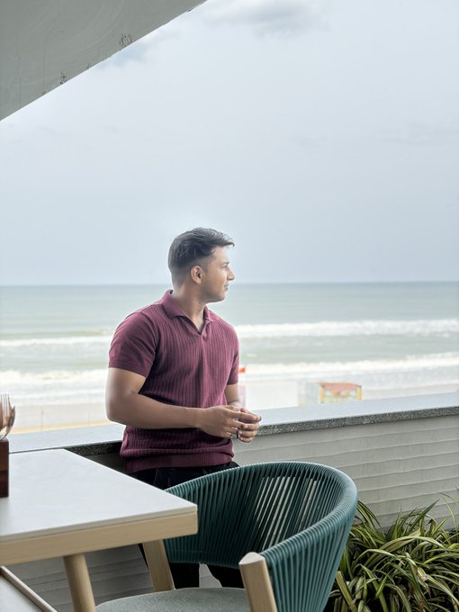 A man in a maroon polo shirt stands on a balcony looking out at the ocean waves on a cloudy day, with a green wicker chair and table visible in the foreground.