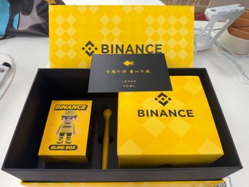 The image features a box containing two items, one of which appears to be a toy or figurine. The other item inside the box is a business card for Binance, a company that specializes in digital currency trading. The card has a yellow background with black text and includes an illustration of a fish on it.