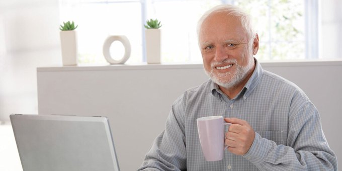A stock photograph of a smiling elderly man with white hair and beard, wearing a blue checkered shirt, holding a lavender mug while seated at a laptop in a bright modern interior.