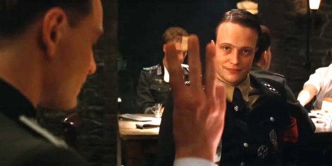 In the image, there are two men dressed in military uniforms sitting at a table with their hands raised. One man has his hand up while the other one has his hand down. The scene appears to be set in a restaurant or dining area where they might be having dinner together. A third person is also present in the background, but only partially visible and not interacting with the main subjects of the image.
