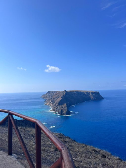 A breathtaking ocean view features a rugged island rising from brilliant turquoise waters under a clear blue sky, viewed from a cliffside overlook with a wooden railing—perfectly capturing the serene, uplifting spirit of a "Gm familia" greeting.