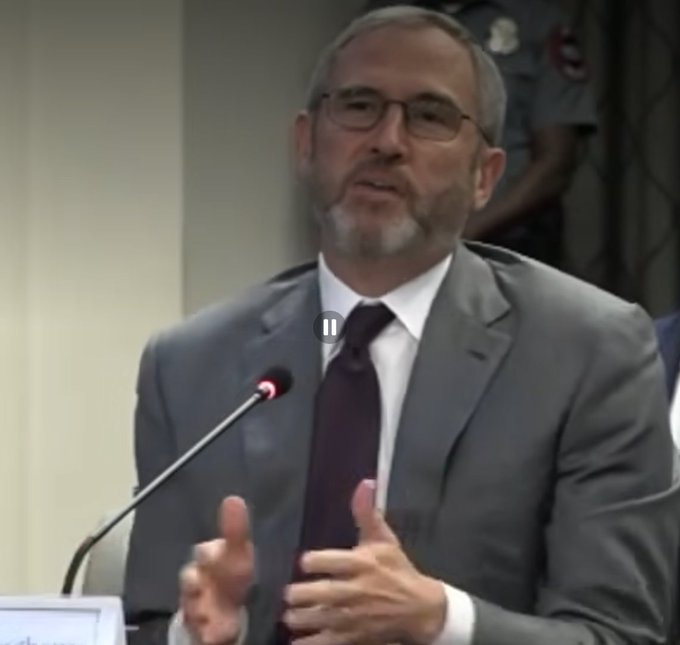 A man with a gray beard and glasses, wearing a gray suit and dark tie, speaks at a microphone during what appears to be a CFTC meeting, gesturing with his hands. The image relates to the post as it captures the moment when Brad Garlinghouse, CEO of Ripple, addresses the panel, highlighting how he and the company were negatively impacted by the prior administration's regulatory stance.
