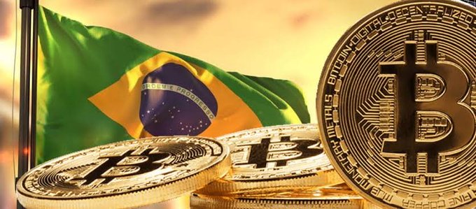 Three golden Bitcoin physical coins displayed in front of the Brazilian flag against a sunset backdrop.