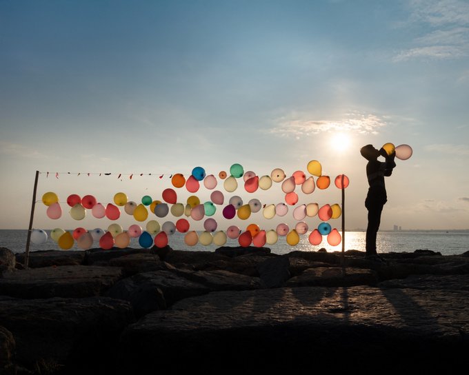 A silhouette of a person standing by a display of colorful balloons strung along a line, backlit by the sun over the ocean on a rocky shoreline.