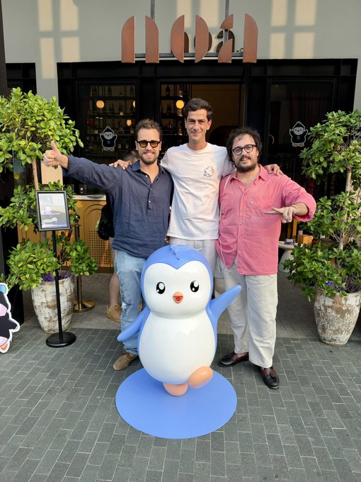 Three cheerful men pose with arms around each other behind a large blue penguin mascot outside what appears to be the "monn" restaurant, with greenery and bar stools visible in the background. The image captures a friendly, team-bonding moment that fits the "Solana huddle" caption, suggesting a casual gathering of the Solana community or team in a sunny, welcoming setting.