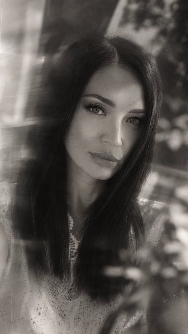A black and white portrait photograph of a young woman with long dark hair, smiling softly and looking at the camera through a window.