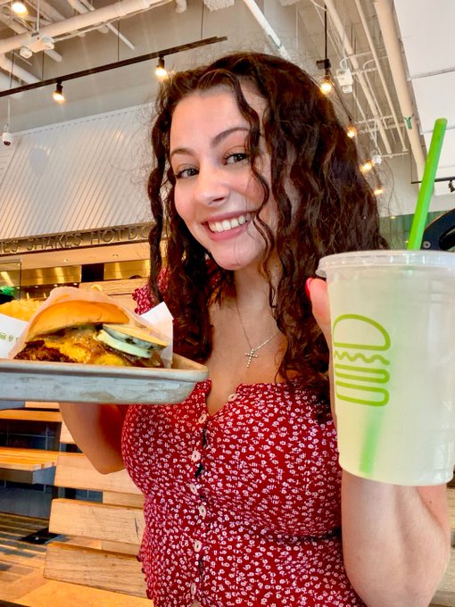 A smiling woman in a red floral dress proudly holds up a cheeseburger on a plate and a lemonade from "C3SU" in celebration of National Cheeseburger Day, showing off her burger pick.