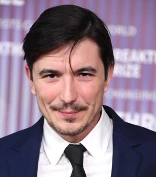 A man in a navy suit, white shirt, and black tie stands smiling in front of a backdrop reading "iHeartRadio," "WORLD," and "BREAKFAST," with the accompanying post connecting him to Robinhood CEO Vlad Tenev. The image serves as a visual reference to Tenev while the caption highlights his comments on the Robinhood blockchain.