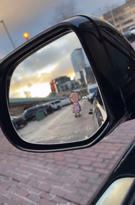 A photograph of a car's side mirror reflecting a city street at sunset, with a cartoon character sticker superimposed on the reflection.