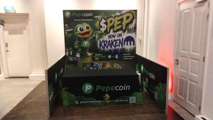 The image features a large advertisement for a game with various graphics, including a cartoon monster that appears as a character from the video game "KrakeEN". The company behind this game, known as "Pepecoin", has sponsored the promotional display. The advertisement is displayed at an event space or convention center, possibly attracting gamers and enthusiasts to try out the game for themselves.