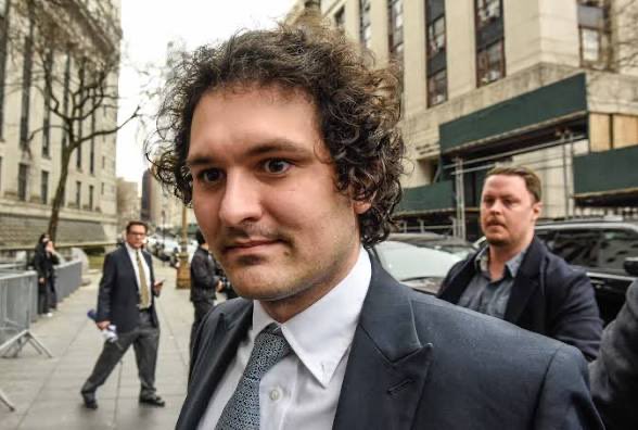 A photograph of a young man with curly hair wearing a suit and tie standing on a city street.