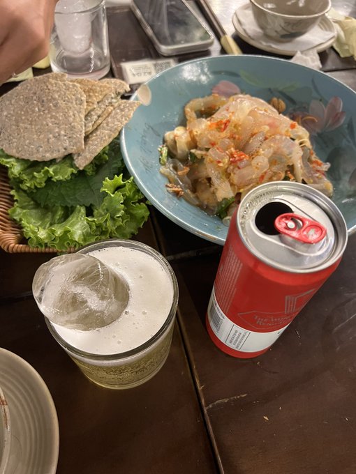 A table at a restaurant with a glass of beer, a red can of beverage, a bowl of raw shrimp or seafood salad with chili, and a basket of flatbread with lettuce.