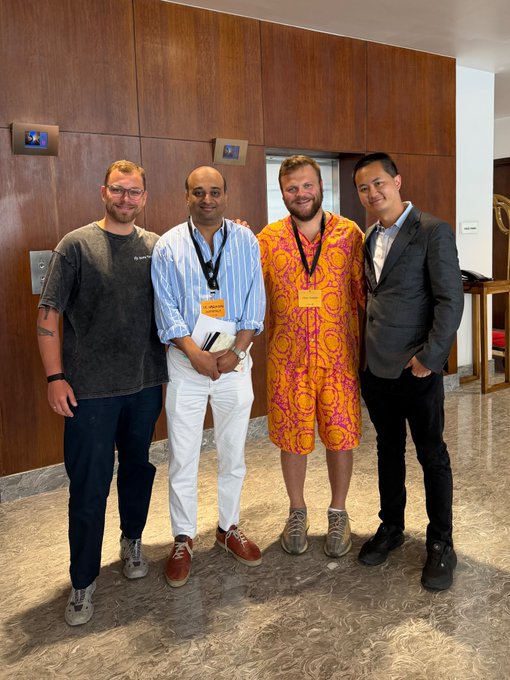 Four men stand side-by-side in an indoor setting with wood-paneled walls and elevator doors, one notably wearing a bright pink and yellow patterned romper.