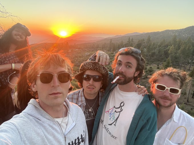 A group of five young people pose for a selfie outdoors at sunset with mountains and a forested landscape in the background; some wear sunglasses, one wears a bucket hat, and one wears a shirt with a logo.