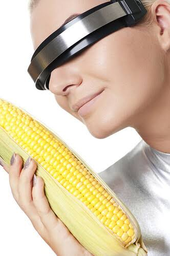 A woman wears a sleek black visor over her eyes while holding an ear of corn close to her face with both hands, playfully obscuring her vision. The image humorously illustrates the post's "leetcorn" pun by pairing the high-tech eyewear with fresh corn, evoking a quirky, sensory experience.