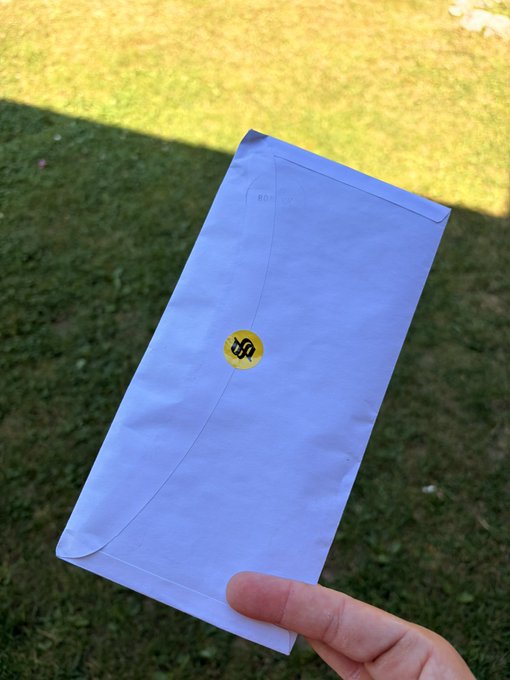 A hand holding a white envelope with a yellow seal on grass, appearing to contain a document.
