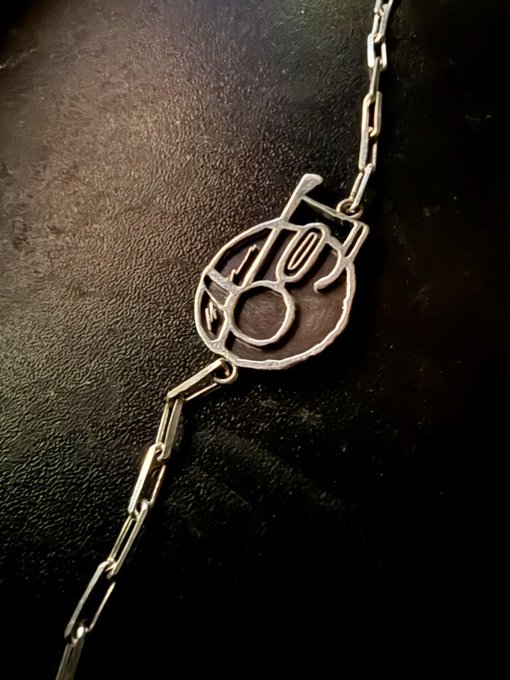 A close-up photograph shows a silver chain bracelet featuring a circular pendant with stylized cutout lettering, displayed on a dark leather surface.