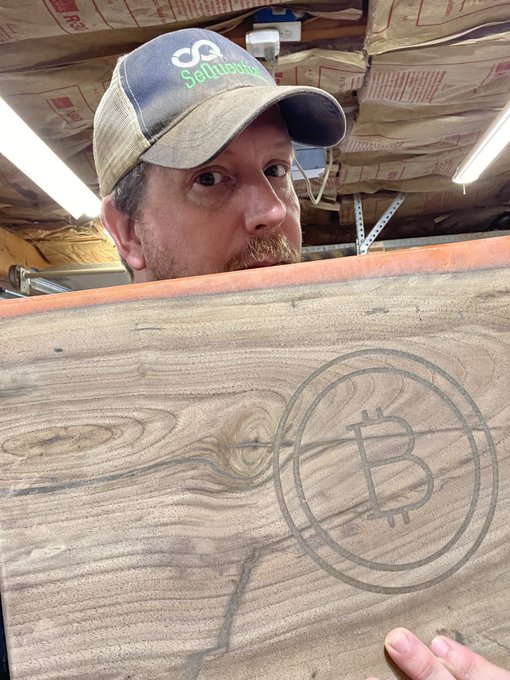 A bearded man wearing a 'Settlerstruck' cap holding up a wooden board with a Bitcoin logo etched into it, standing in what appears to be a basement or workshop.