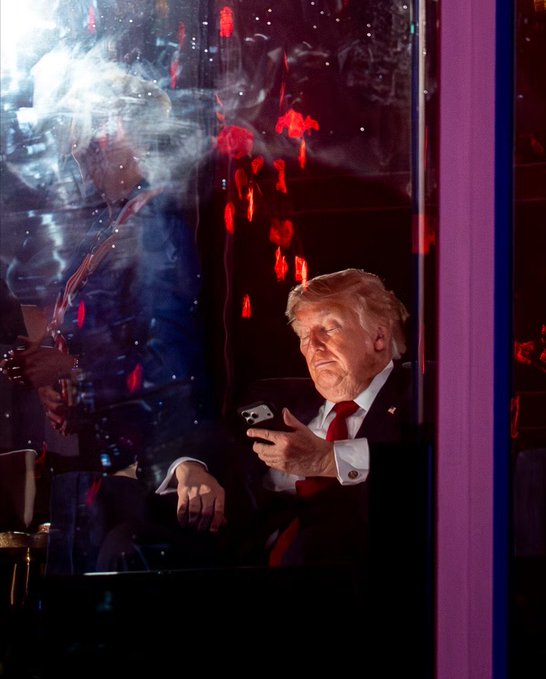 A man in a suit looking at his phone, viewed through a reflective glass surface with colorful light reflections.