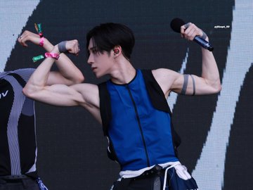 A performer with short dark hair in a blue and black sleeveless outfit holds a microphone on stage while flexing his arm, with a wristband visible.