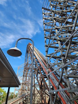 A wooden roller coaster structure rises against a blue sky with scattered clouds, with a vintage street lamp visible in the foreground.