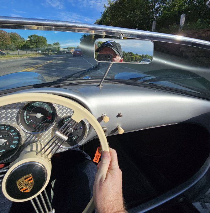 A first-person view from inside a vintage Porsche shows the driver's hand on the cream-colored steering wheel, with the rearview mirror reflecting a person taking the photo.