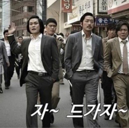 A group of men in suits walk down a street with Korean shop signs in the background, with white Korean text "자~ 드가자~" overlaid at the bottom.