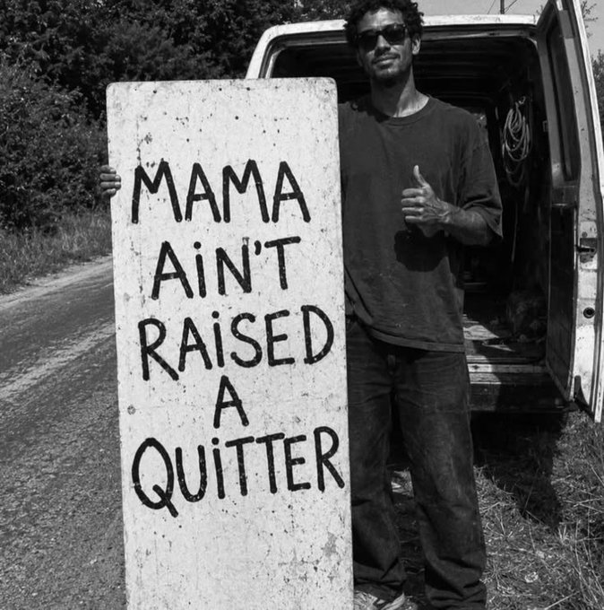 A black-and-white photo of a man in sunglasses standing next to a van, holding a handwritten sign that reads 'MAMA AIN'T RAISED A QUITTER'.