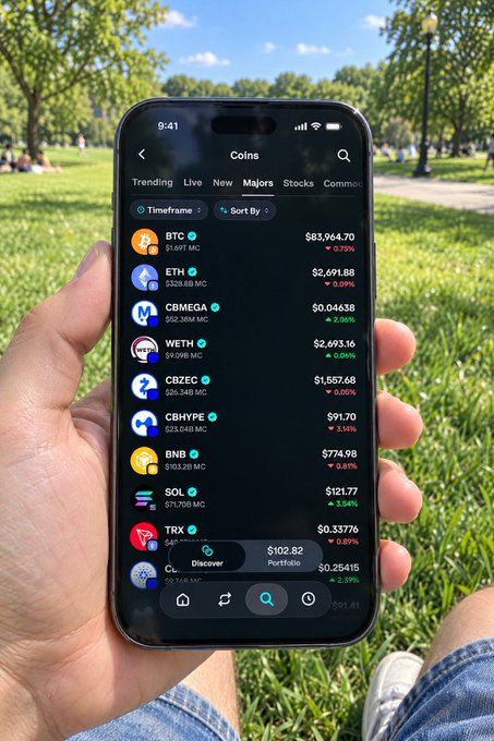 A hand holds a smartphone in a sunny park, displaying a crypto tracking app with real-time prices for BTC ($83,964.70), ETH ($2,691.88), SOL ($121.77), and several altcoins. The relaxed outdoor setting perfectly matches the caption's sentiment of casually checking market charts while enjoying a nice walk.