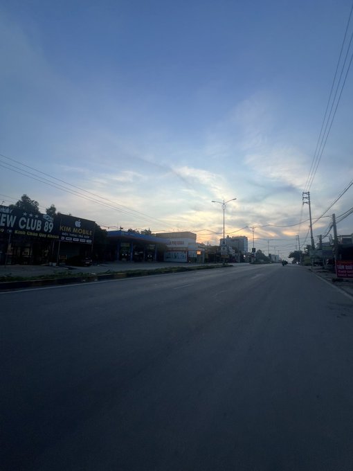 A view down a wide, empty street at dusk or dawn, featuring businesses like 'CREW CLUB 69' and 'KIM MOBILE' on the left side under a partly cloudy sky.