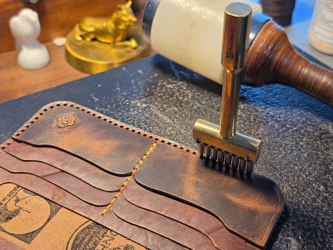 A close-up photograph of leatherworking tools, including a stitching chisel with multiple prongs resting on a piece of curved brown leather. A mallet and other crafting items are visible in the blurred background.