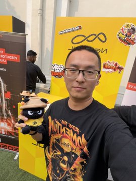 A man wearing glasses and a 'Mutilator' t-shirt posing at what appears to be a gaming/event booth with yellow signage, holding a small tiger plush toy.