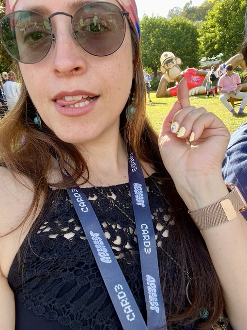 A woman wearing round sunglasses and a red headscarf takes a selfie at an outdoor festival, wearing lanyards labeled "CARDI B," "HIPGNOSIS," and "CRYPTO," while pointing toward an inflatable monkey mascot behind her, playfully riffing on the "GM Apes" greeting by combining crypto/Web3 culture with a literal monkey at what appears to be an ApeFest-style event.