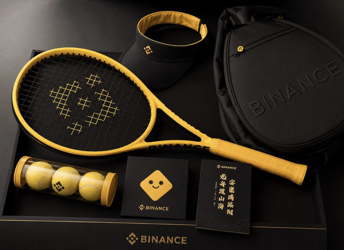 The image features a tennis racket, balls, and other sports equipment displayed together with a caption that reads "One of the most popular sports brands in the world, Binance, has released their new line of tennis rackets". The items are arranged on a black surface, creating an appealing visual presentation.

The main focus is on the tennis racket, which is prominently placed at the center of the image. Surrounding it are several tennis balls and other sports equipment such as a backpack and a hat. The caption provides context about the brand "Binance" and their new line of tennis rackets.