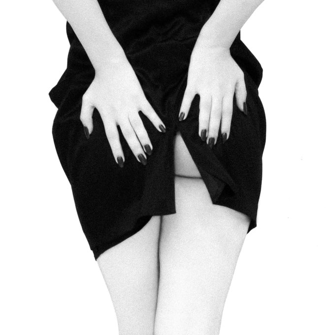 A black-and-white close-up photo showing a person's hands resting on their black skirt, with dark-painted nails visible and pale legs crossed below.