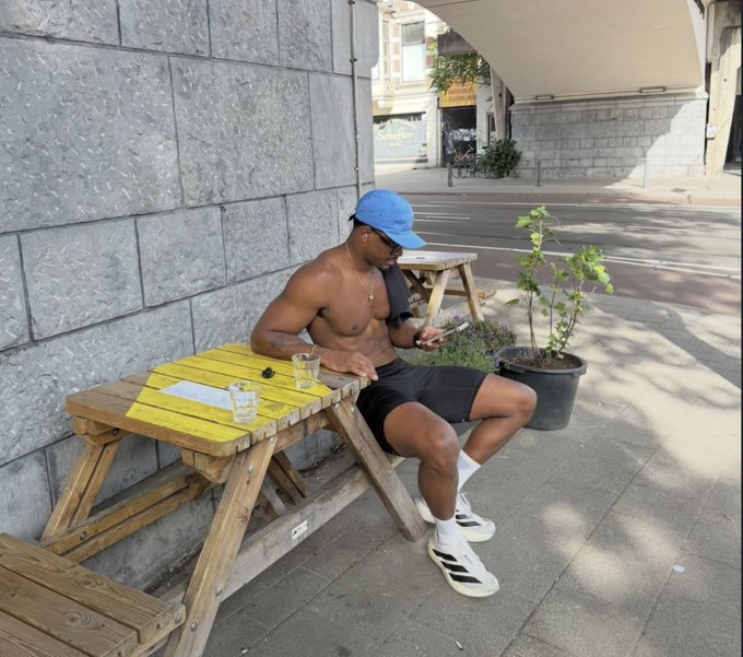 A shirtless man in a blue cap and shorts sits at a yellow picnic table checking his phone, embodying a relaxed "GM" (good morning) vibe as he enjoys the sunny outdoor setting. The post's call to "level up today" pairs with the image's laid-back street-side scene, inviting others to join in or engage.