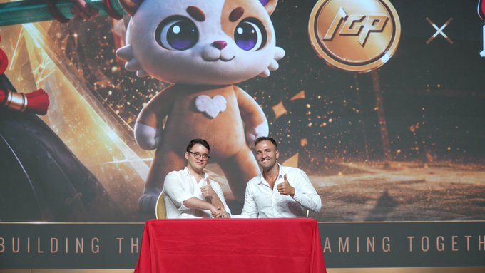 Two men in white shirts give thumbs-up gestures while seated at a red table in front of a large promotional backdrop featuring a cartoon mascot and an 'ICP' logo, with partial text reading 'BUILDING TH' and 'AMING TOGETH'.